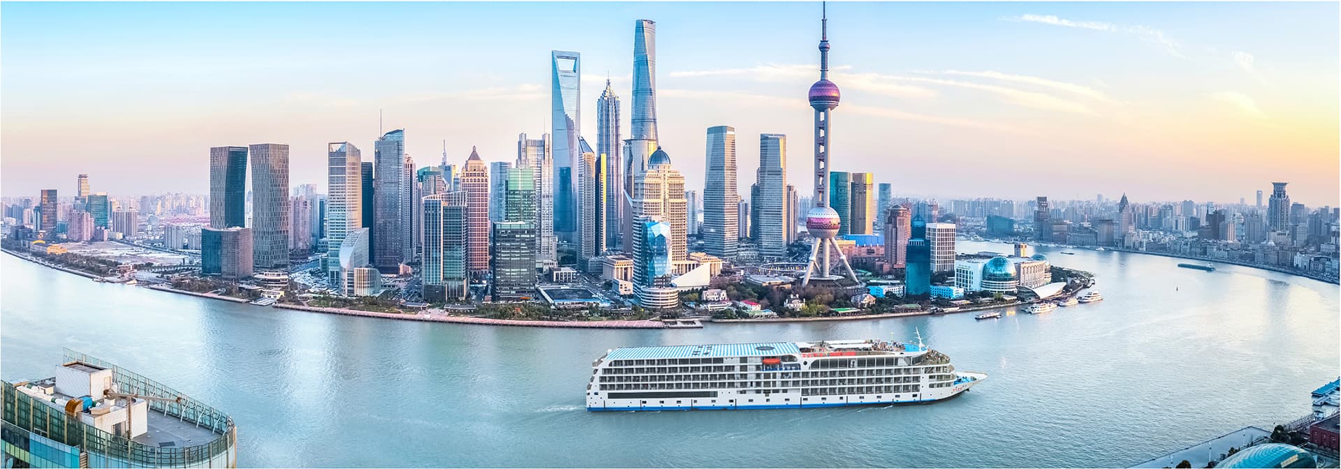 A river cruise vessel on a broad river below a modern city skyline at dusk