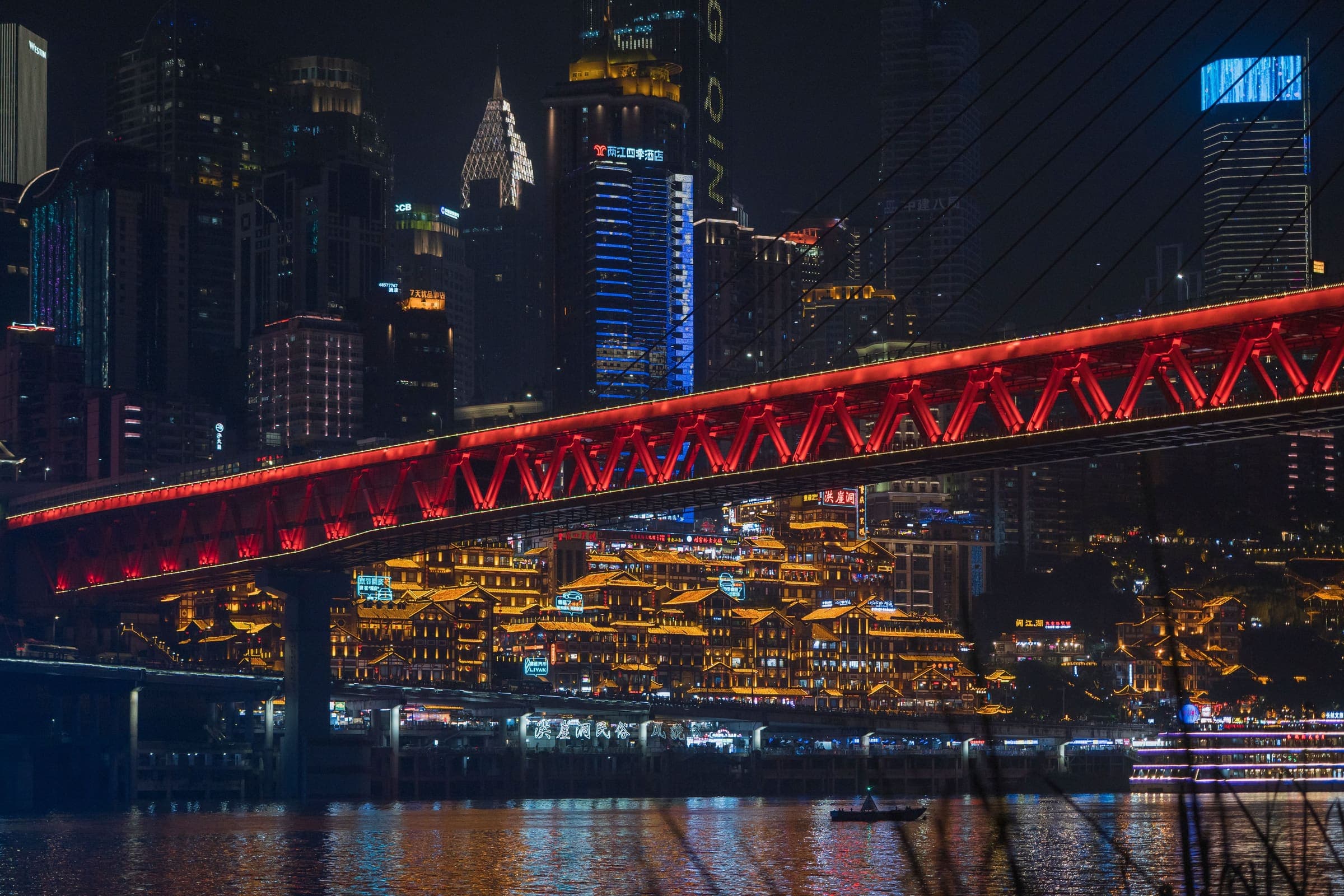 Chongqing after dark from the river — a lit red bridge span, tiered lantern-lit buildings stacked on the bank beneath it, and the city towers behind