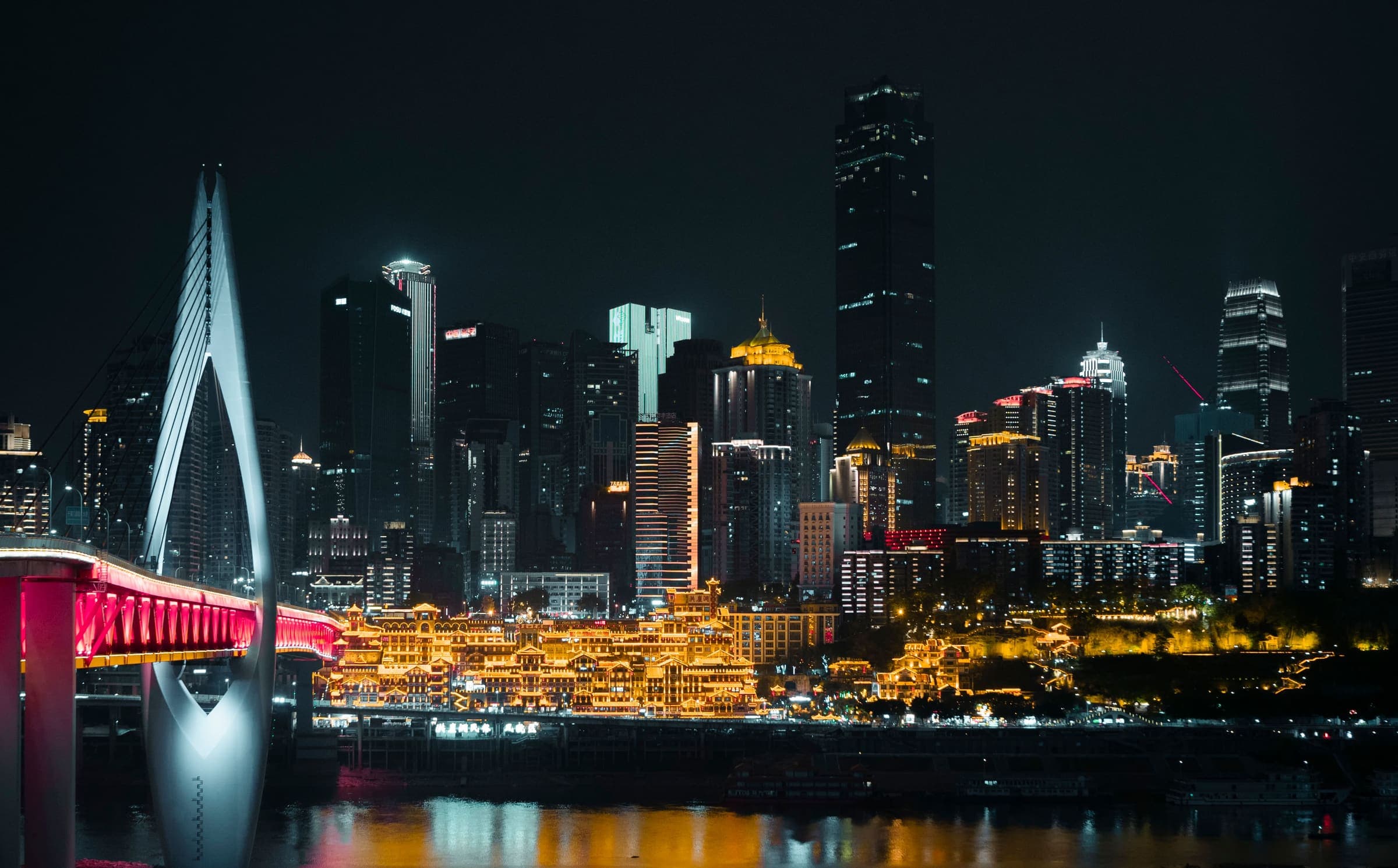 The Chongqing skyline after dark, its towers and lit bridge reflected in the river